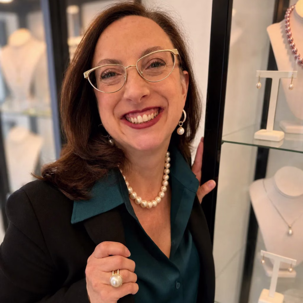 Trish Arato standing beside a jewellery display while wearing a pearl necklace, earrings, and ring.